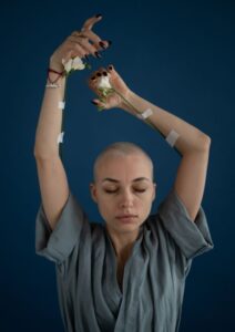 Femme au crâne rasé ayant les bras levés avec une fleur scotchée à chaque bras représentant les tuyaux utilisés pour soigner du cancer